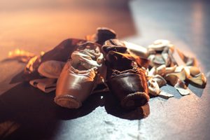 A pair of tan and a pair of brown ballet pointe shoes lie side by side on a stage floor, surrounded by light from above, with their ribbons scattered around them.