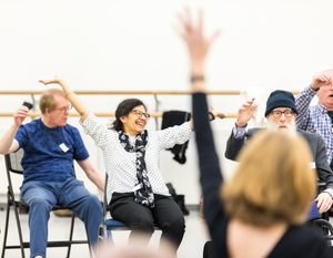 A group of adults sit in chairs in a bright room, smiling and raising their arms enthusiastically during what appears to be a group activity or exercise. One person in the foreground raises an arm.