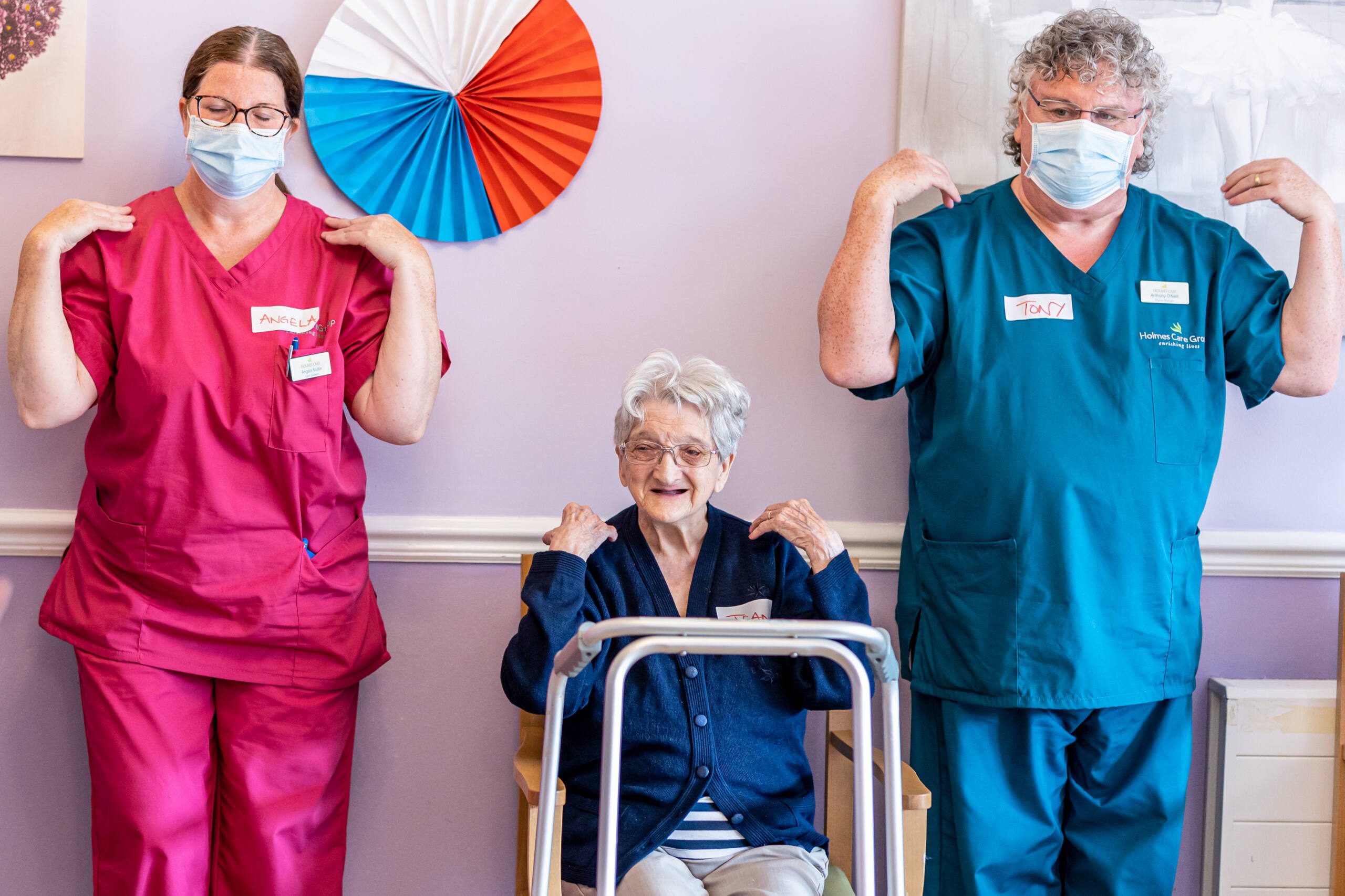 Two healthcare workers in scrubs and masks stand on either side of an older woman seated with a walker, all smiling and raising their arms in a dance against a pastel-colored wall.