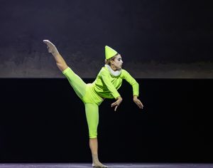 A dancer in a bright green costume and pointed hat balances on one leg with the other leg extended high behind her on a dark stage.