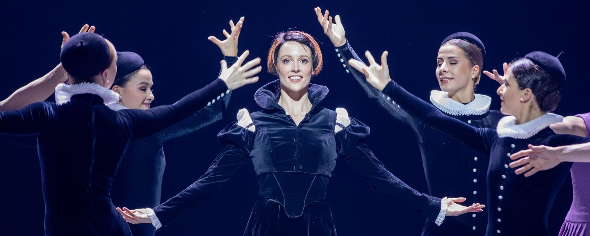 A woman in a black Tudor-style dress stands center stage, surrounded by four dancers in black outfits with white ruffs, their arms extended toward her against a dark background.