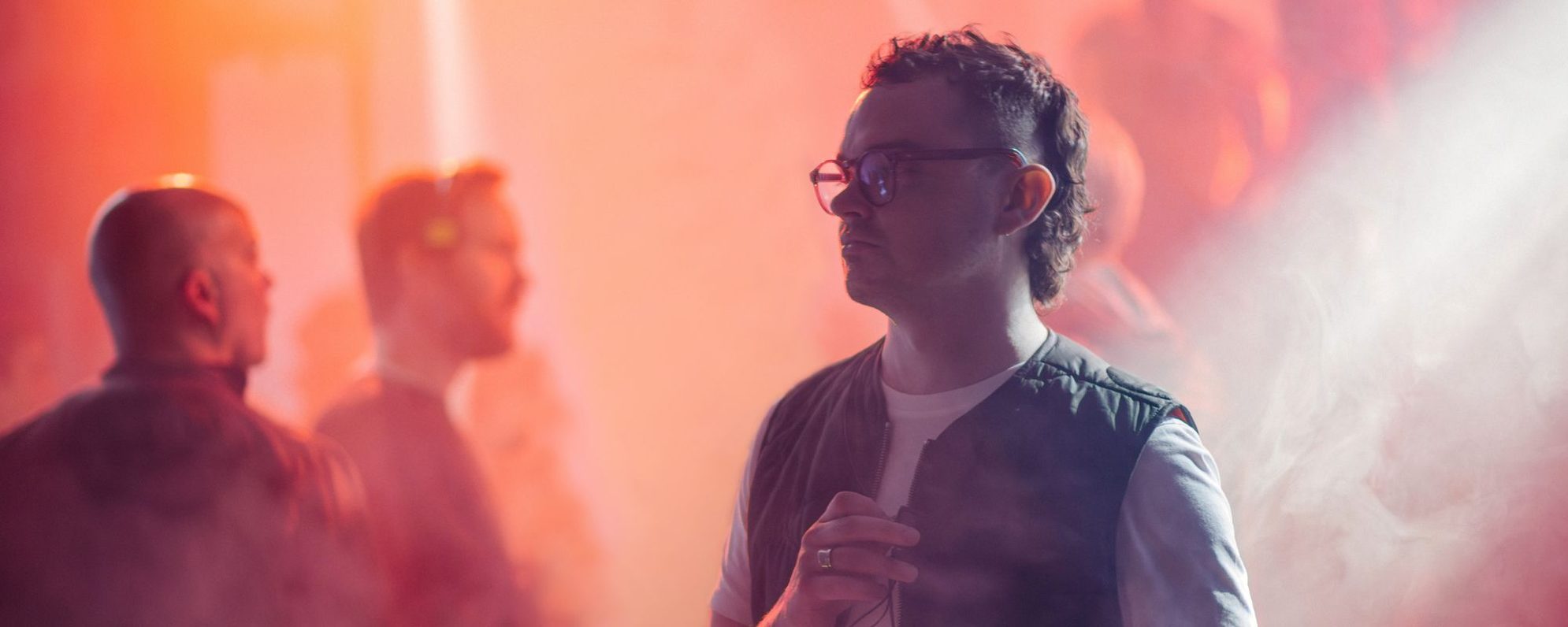 A man wearing glasses and a vest stands in a smoky, red-lit room with blurred figures in the background, creating a moody and atmospheric scene.