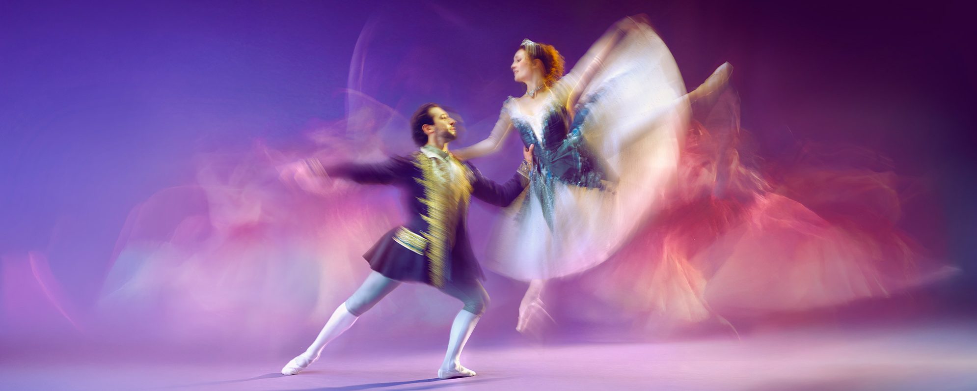Two ballet dancers perform gracefully on stage. The male dancer extends his arms while the female dancer leaps mid-air, her tutu and movement creating a blurred, colourful effect against a purple background.