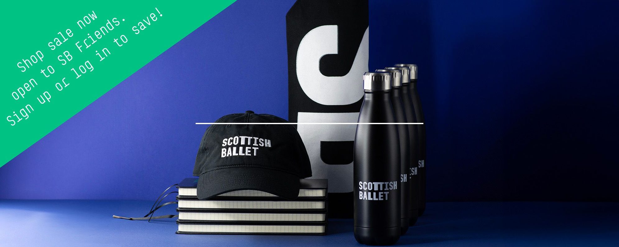A black cap labeled Scottish Ballet, four black water bottles, and stacked notebooks are displayed on a blue background. A green banner in the corner reads, Shop sale now, open to SB friends: Sign up or log in to save!