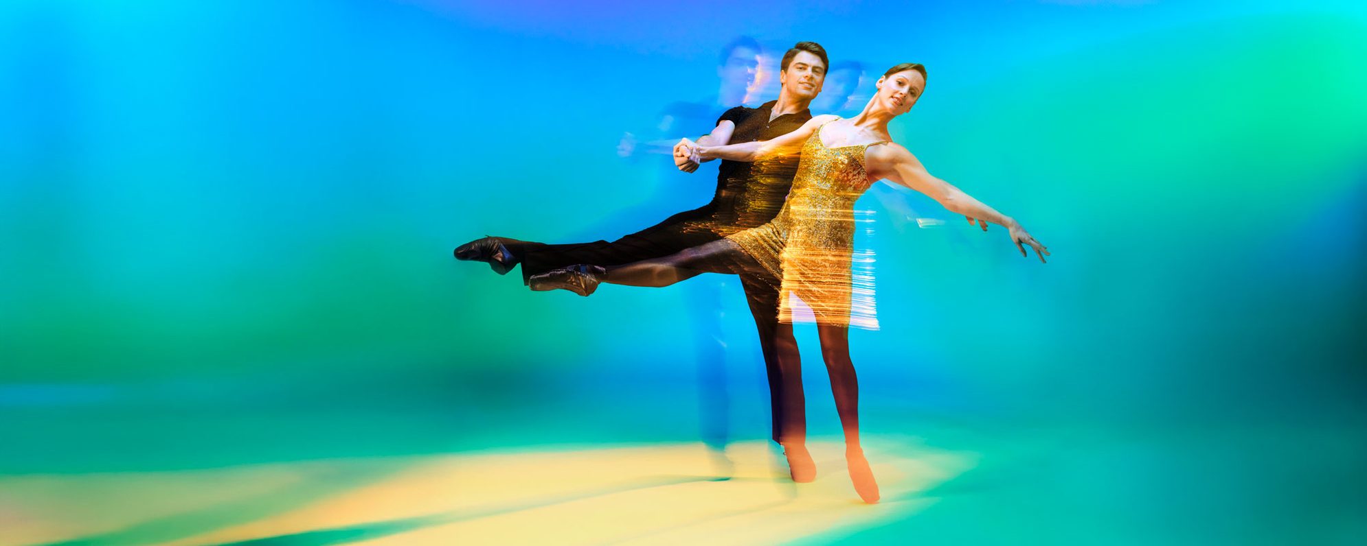 A man and woman perform a dance in dynamic motion, creating a blurred effect. The woman wears a sparkling gold dress and extends her leg, while the man supports her. The background is a gradient of blue, green, and yellow.