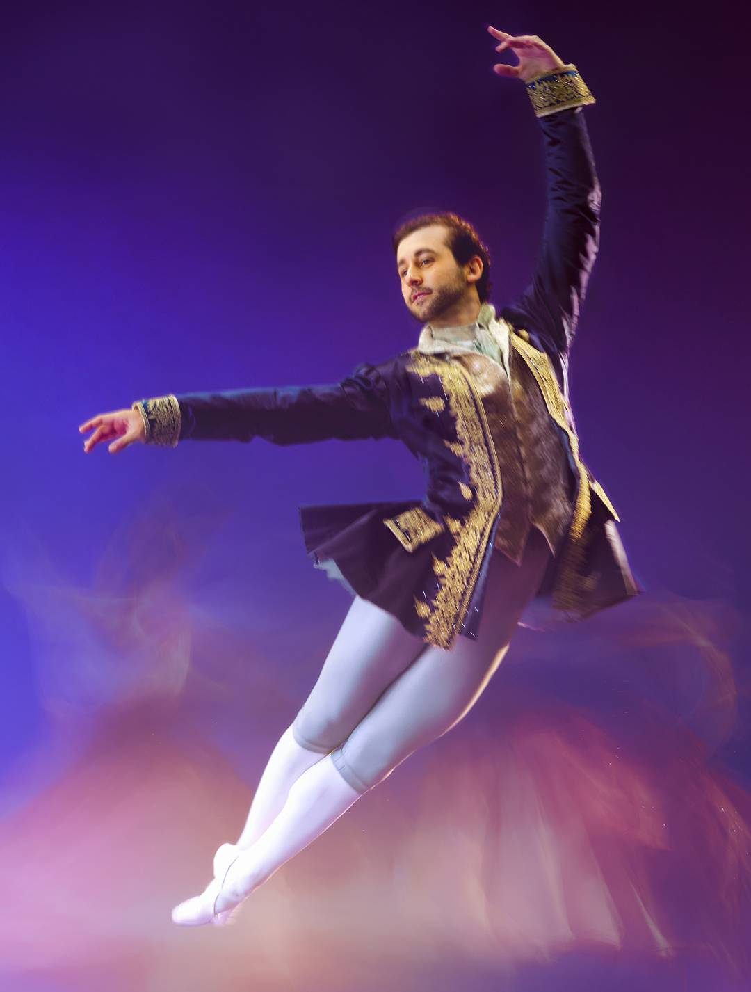 A ballet dancer in an ornate costume leaps gracefully on stage against a blurred, colorful background, capturing motion and elegance.