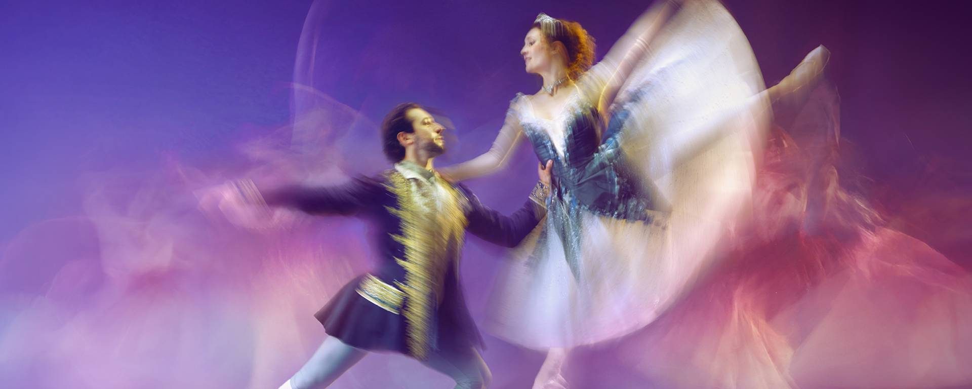 A ballet dancer in a dress and a ballet dancer in a regal coat perform , captured in mid-movement with blurred, flowing motion. The dancer in a dress leaps gracefully , the dancer in a coat supports, both surrounded by a purple-lit background.