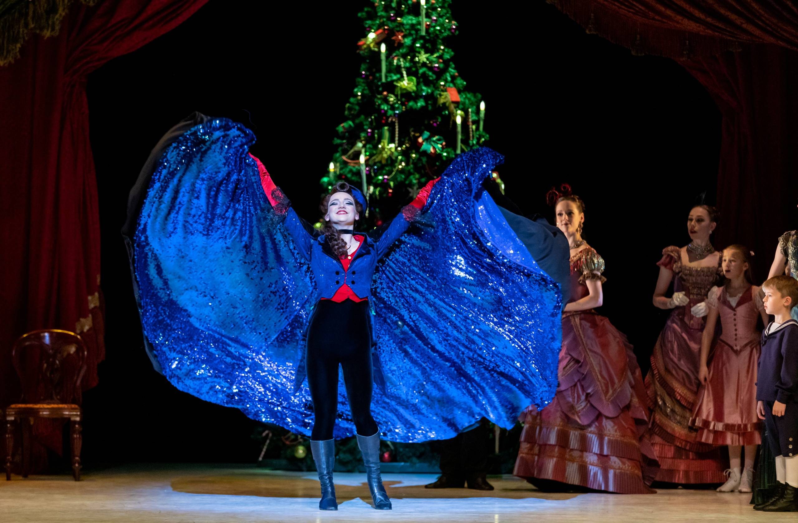 A performer in a sparkling blue cape stands center stage with arms raised, in front of a decorated Christmas tree, while dancers in elegant costumes and children watch in the background.