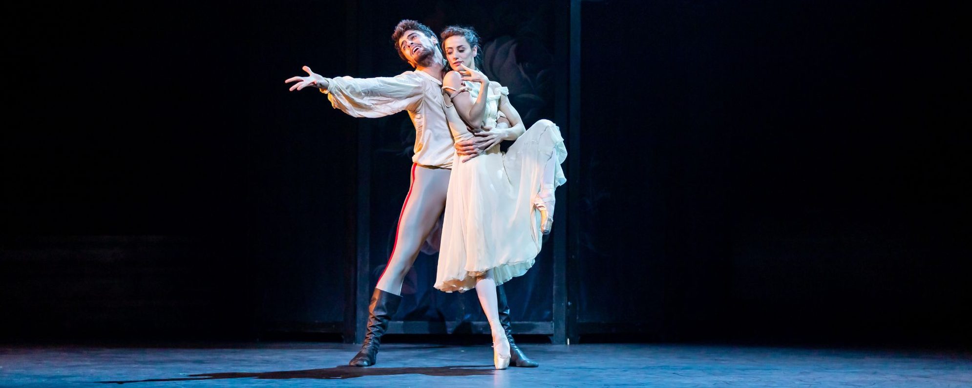 A male and female ballet dancer perform on stage; the man wears a white shirt and boots and holds the woman, who wears a flowing white dress and balances gracefully on one pointe shoe.