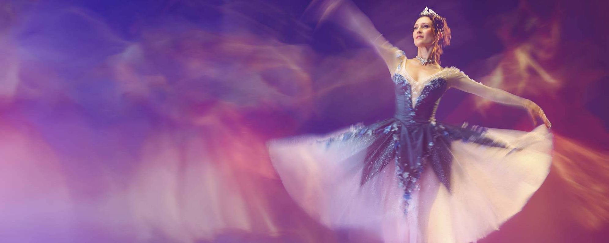 A ballerina in a blue and white tutu dances gracefully on pointe, with one arm raised and motion blur around her. The background is a dreamy blend of pink, purple, and yellow hues.