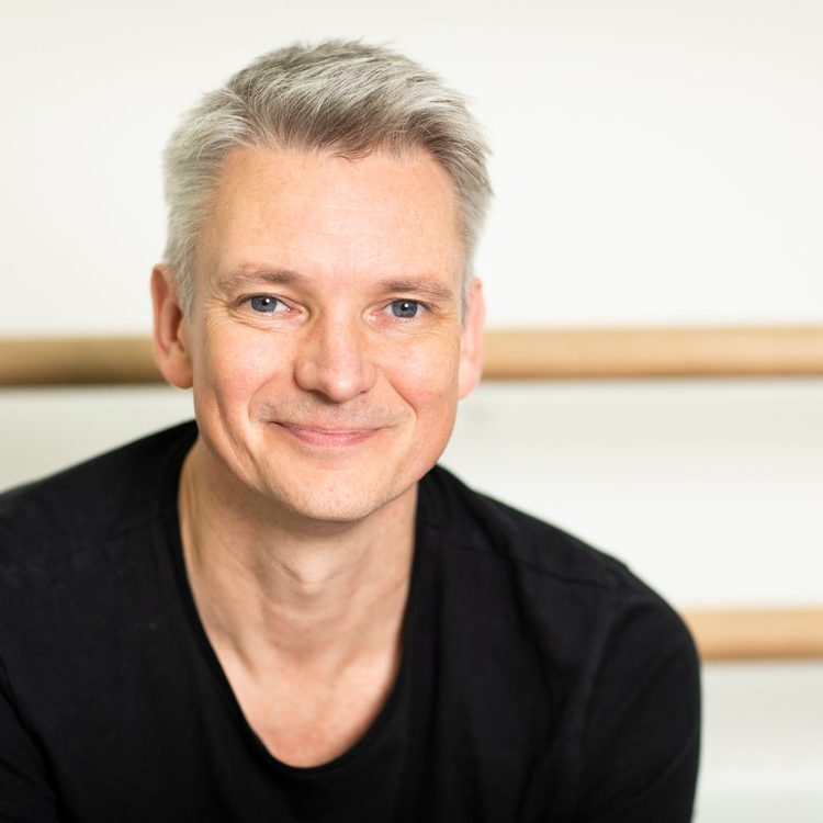 A middle-aged man with short grey hair and a friendly smile is wearing a black shirt. He is sitting in front of a plain background with wooden ballet barres.