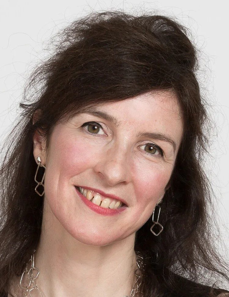 A woman with long dark hair smiles at the camera. She is wearing geometric silver earrings and a matching necklace. The background is plain white.