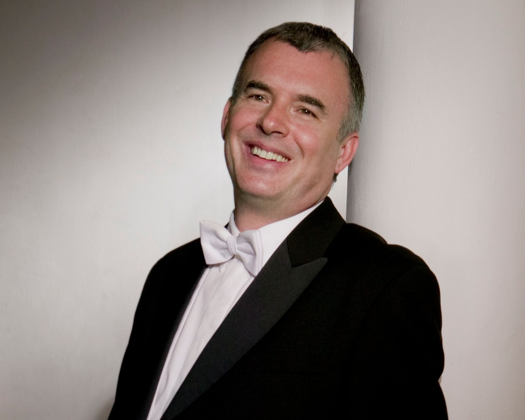 A man in a dinner jacket with a white bow tie is smiling whilst leaning against a light-coloured wall.