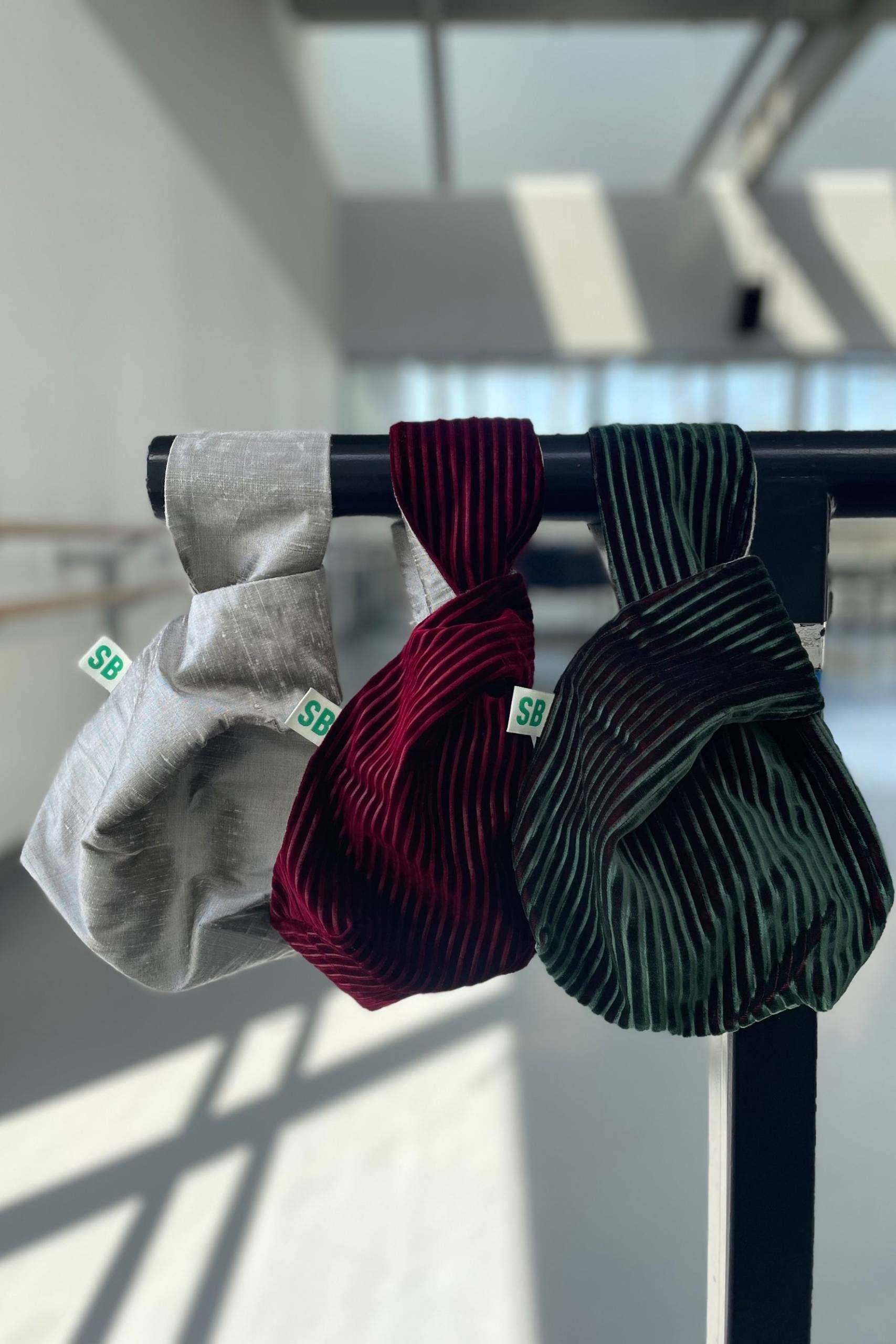 Three small fabric bags—one grey, one burgundy, and one green with black stripes—hang side by side from a black rod in a sunlit room. Each bag has a small white tag labelled “SB.”.