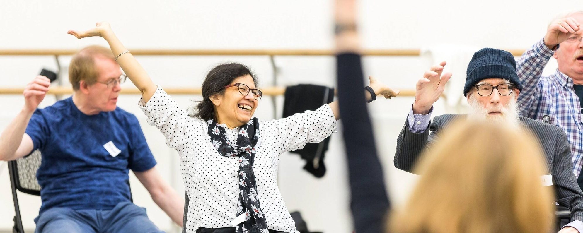 Older adults seated on chairs in a dance studio raise their arms and smile during a joyful movement or dance class, with ballet barres visible on the wall behind them.