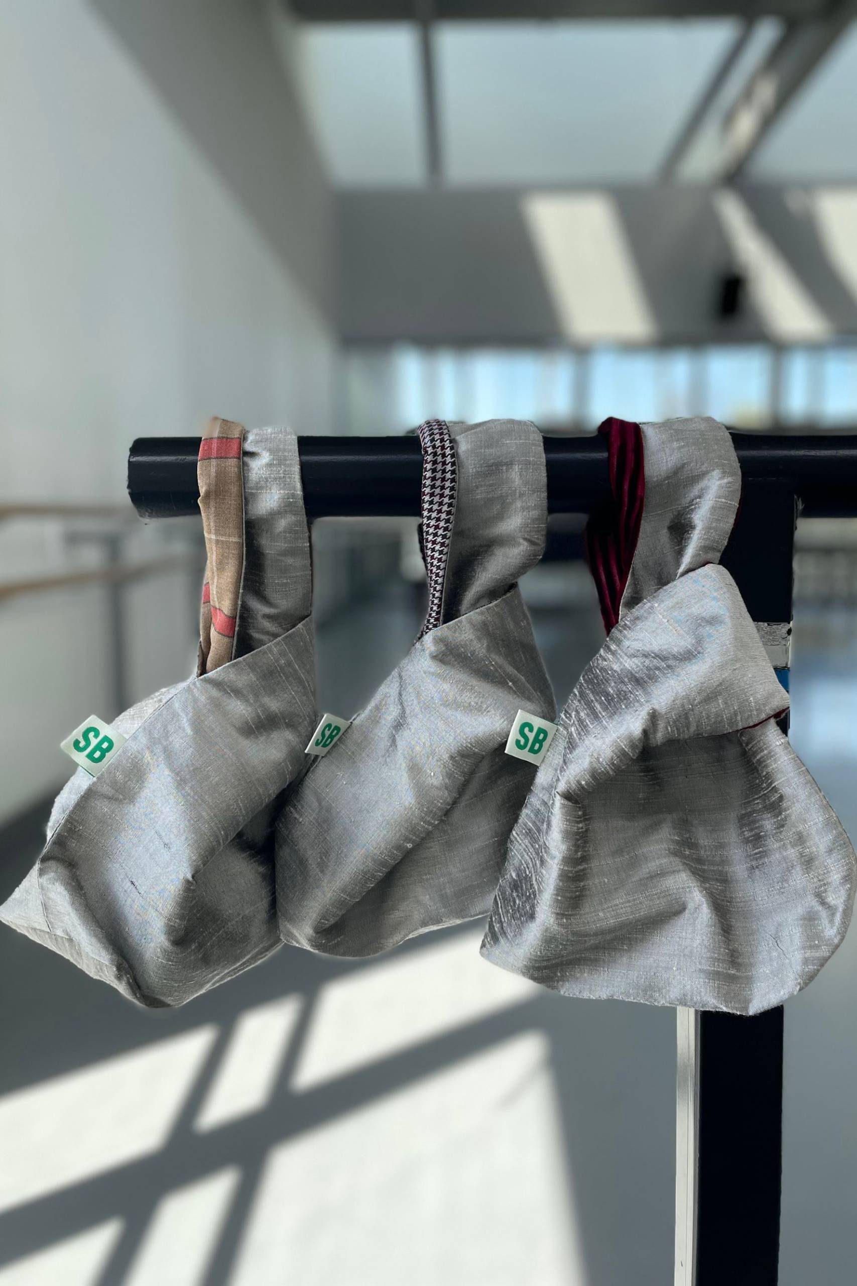 Three small, silver-coloured fabric bags with short handles hang from a black rail. Each bag has a green SB tag, and sunlight streams in from large windows, casting shadows on the floor.