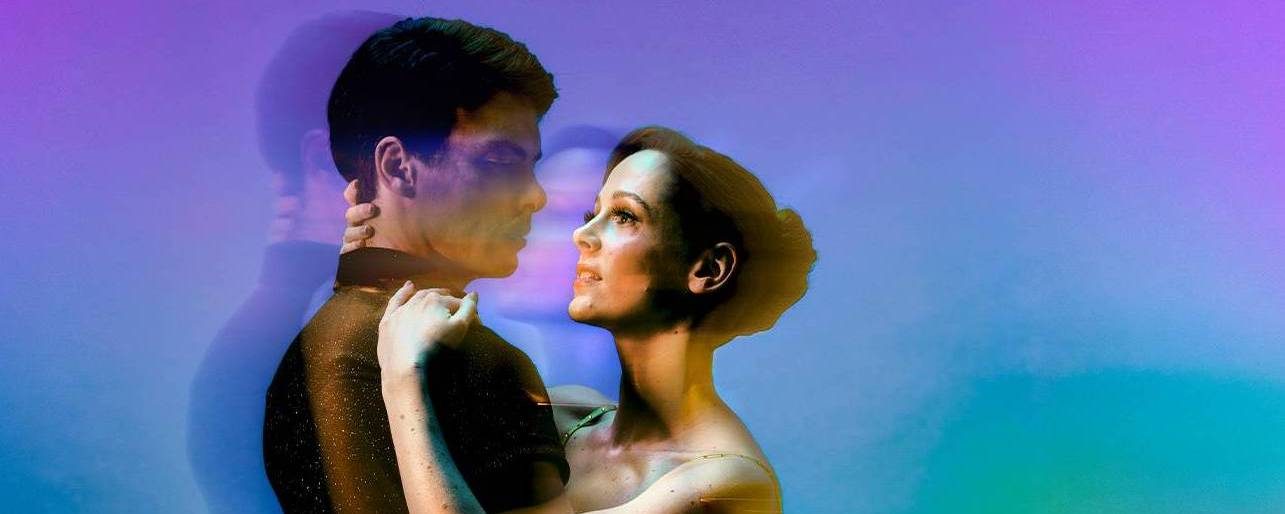 A man and woman in formal attire embrace and gaze into each other’s eyes, standing against a vibrant, colourful, and dreamy background with a soft double-exposure effect.