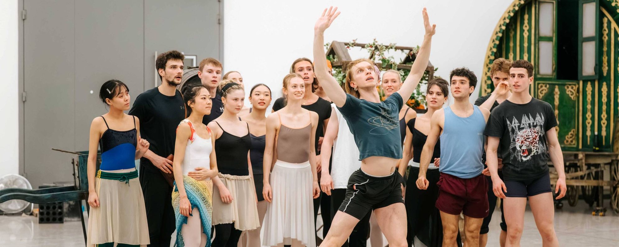 A group of ballet dancers in rehearsal attire watch as one male dancer leaps gracefully with arms raised in front of them in a spacious studio with a large green decorative cart in the background.