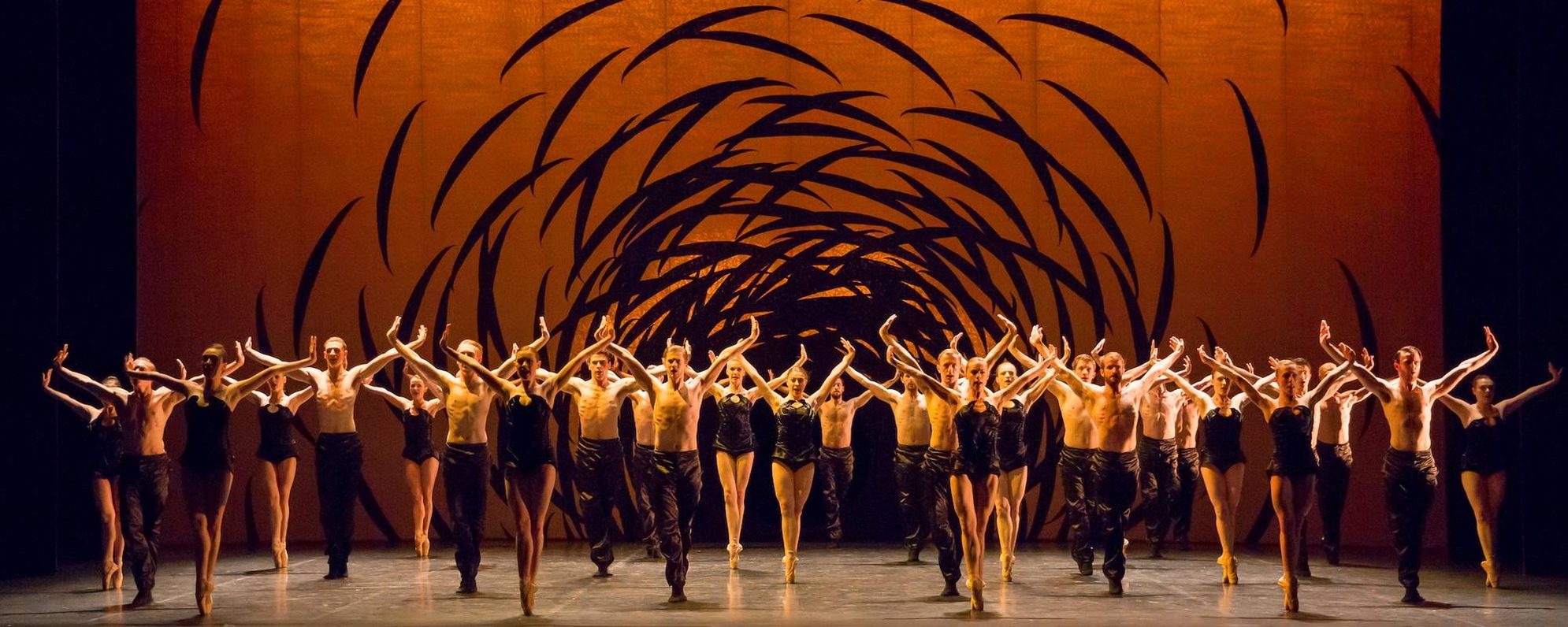 A large group of dancers perform on stage in coordinated formation, arms raised, against an orange backdrop with swirling black shapes, creating a dynamic and energetic atmosphere.