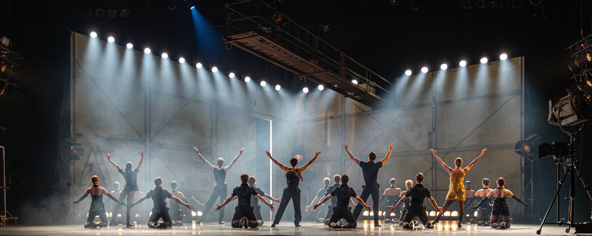 A group of performers on stage, mostly kneeling with arms raised in a V shape, are illuminated by dramatic stage lights against a smoky, industrial-style backdrop.
