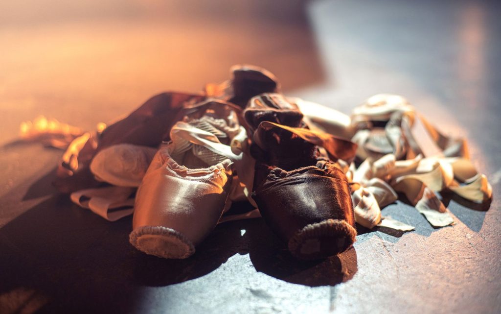 A pile of well-worn ballet pointe shoes in various skin tones resting on a dimly lit floor, illuminated warmly from the side.