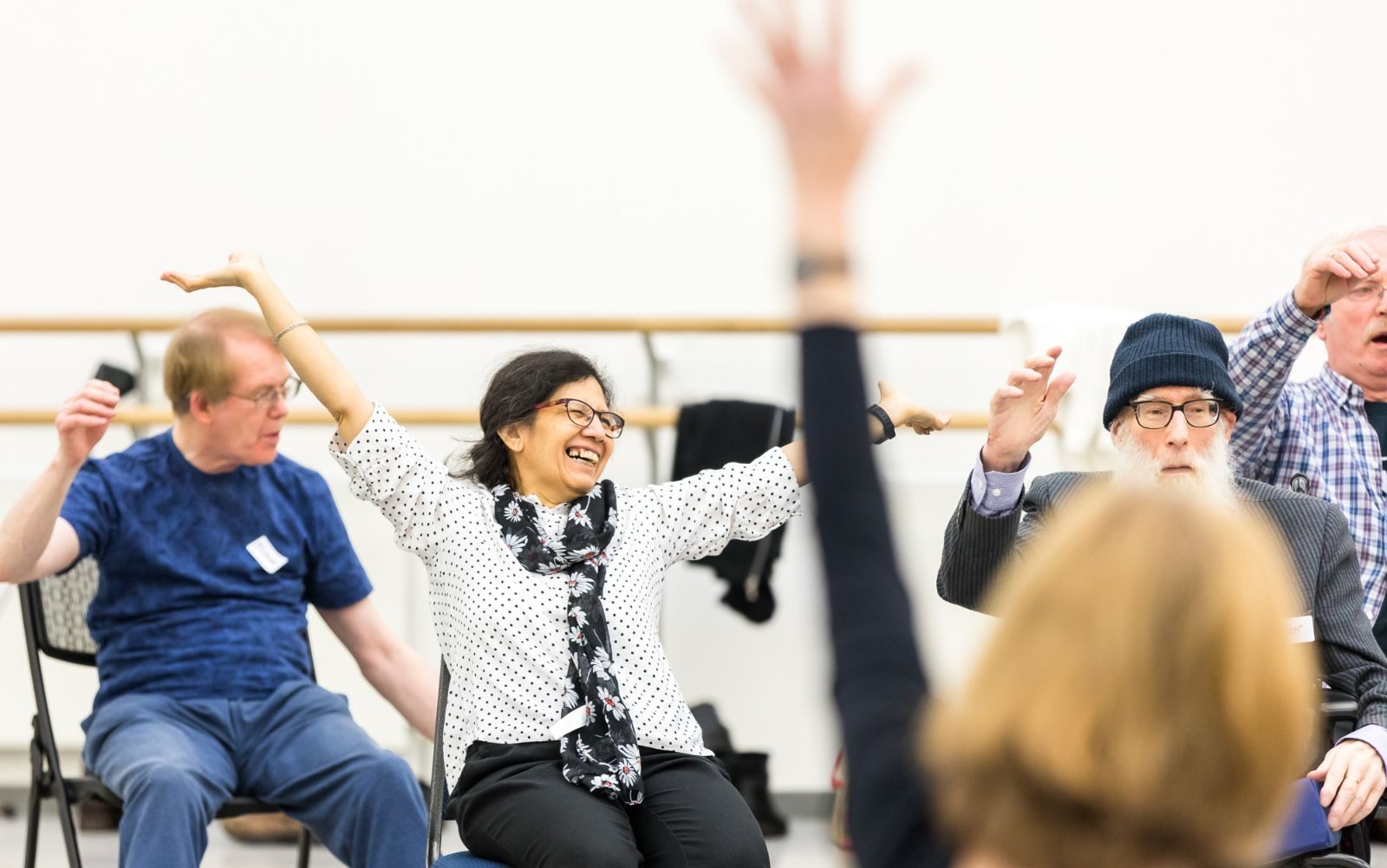 A group of older adults sitting in chairs, joyfully raising their arms in the air during an indoor group activity; one woman in the centre is smiling widely.