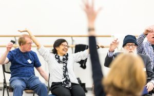 A group of older adults sitting in chairs, joyfully raising their arms in the air during an indoor group activity; one woman in the centre is smiling widely.