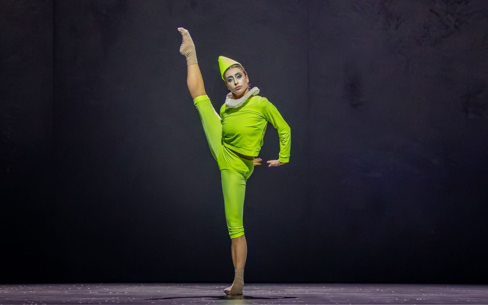 A dancer in a bright green costume and pointed hat performs on stage, balancing on one leg with the other leg extended high, demonstrating flexibility and balance against a dark background.