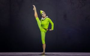 A dancer in a bright green costume and pointed hat performs on stage, balancing on one leg with the other leg extended high, demonstrating flexibility and balance against a dark background.