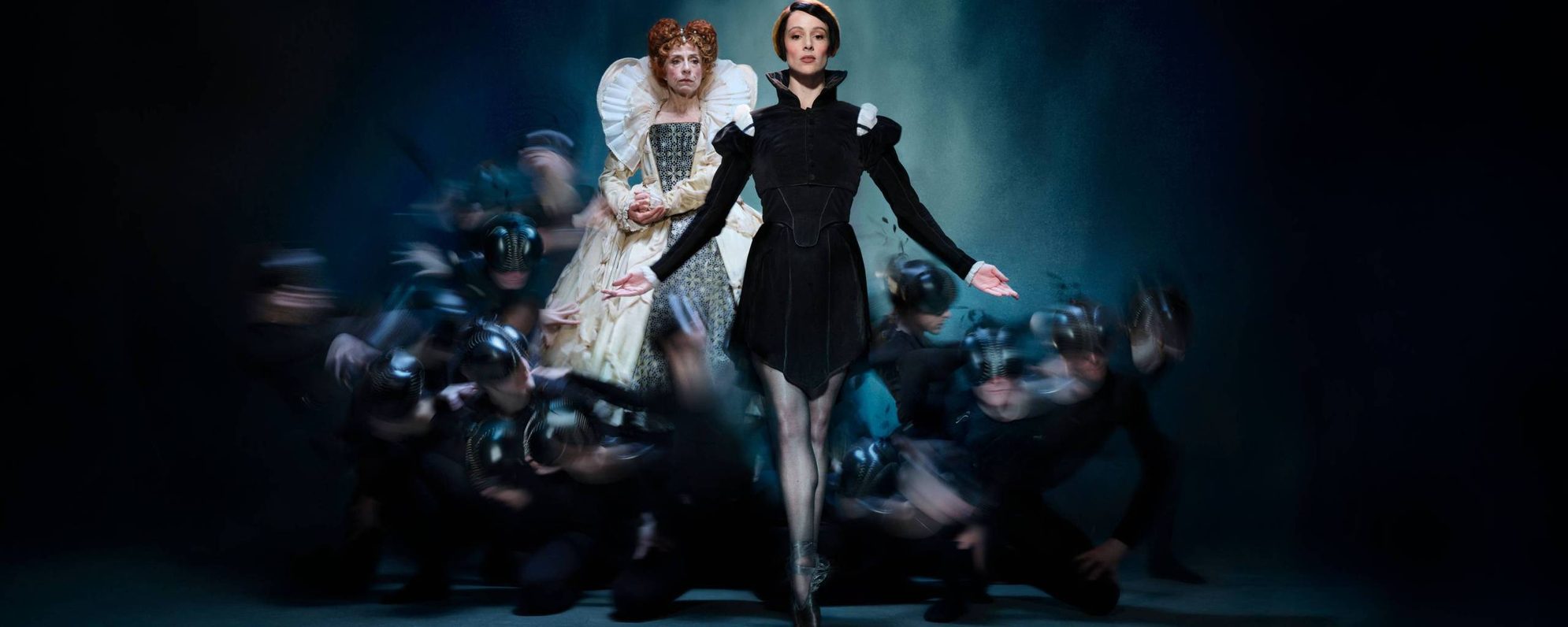 A ballerina in a black outfit poses on pointe centre stage, arms gracefully extended, while a woman in an ornate white dress and ruff stands behind her. Blurred dancers in black crouch around them against a dark background.