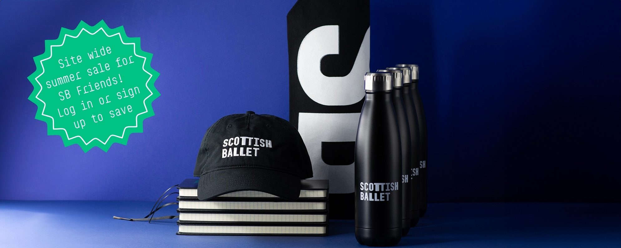 Scottish Ballet merchandise including a cap, water bottles, notebooks, and a tote bag on a blue background. A green starburst label reads, Site-wide Summer Sale for SB Friends! Log in or sign up to save.