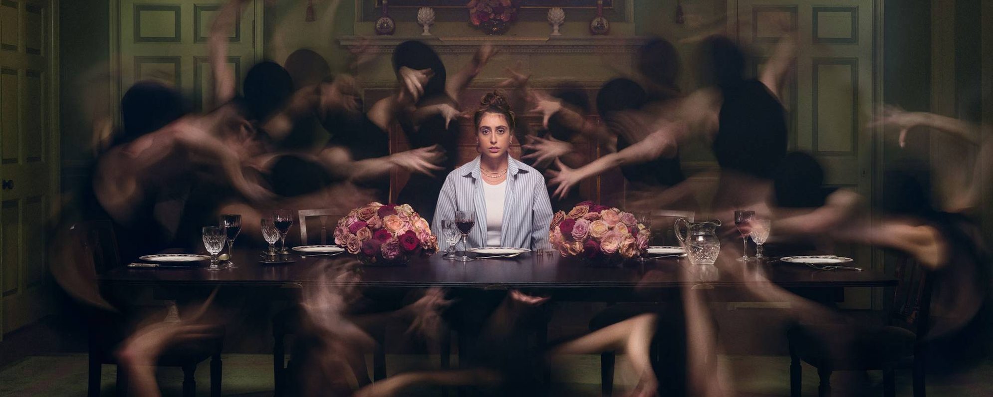 Juliet sits alone at a formal dining table with flowers, surrounded by ghostly, blurred figures in black moving around them, creating a surreal and eerie atmosphere in a dimly lit room.