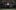 Juliet sits alone at a formal dining table with flowers, surrounded by ghostly, blurred figures in black moving around them, creating a surreal and eerie atmosphere in a dimly lit room.