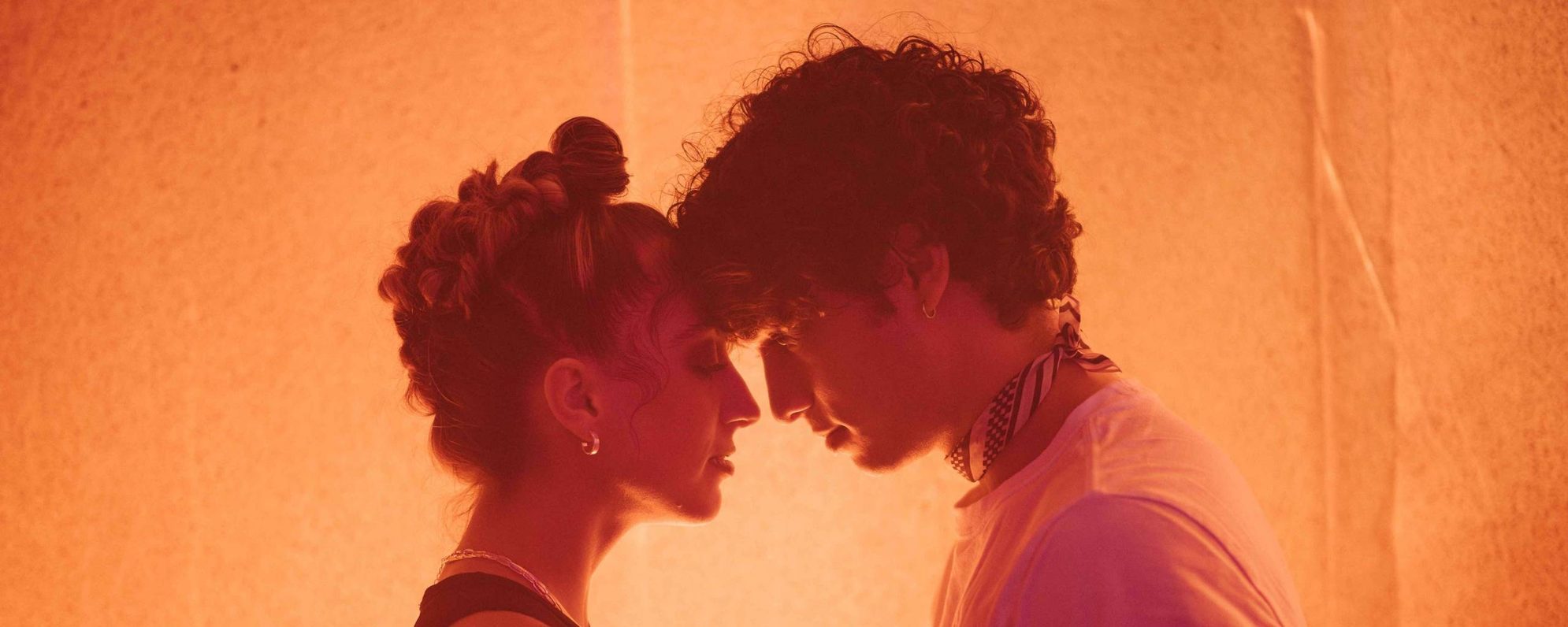 Two people stand close together in warm orange lighting, holding hands and touching foreheads, creating an intimate and tender moment.