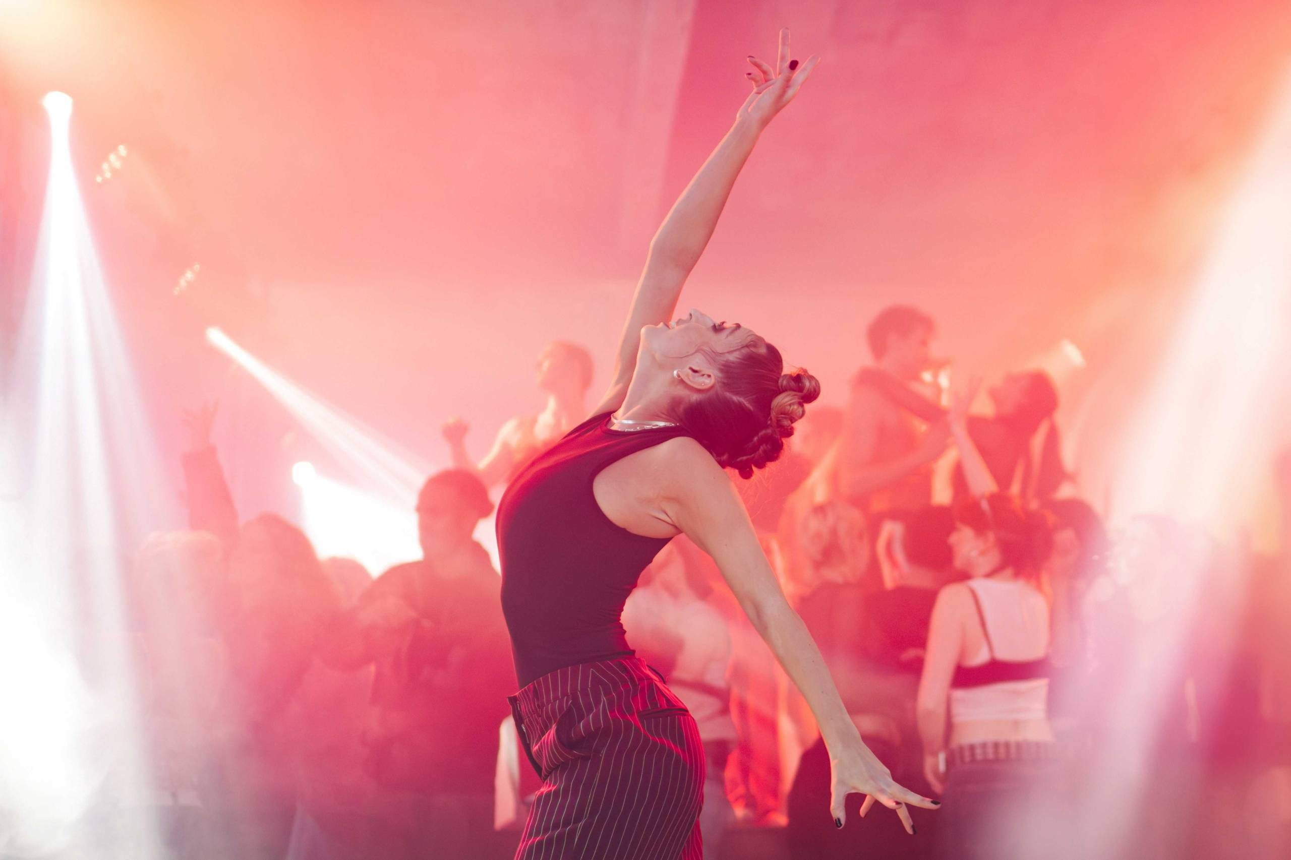 A person in a black top and striped trousers dances with arms outstretched, surrounded by colourful lights and a crowd of people in a lively, energetic atmosphere.