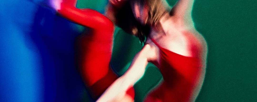 Two dancers in red leotards pose gracefully against a green and blue background, their movements captured with a blur effect that creates dynamic, abstract shapes.