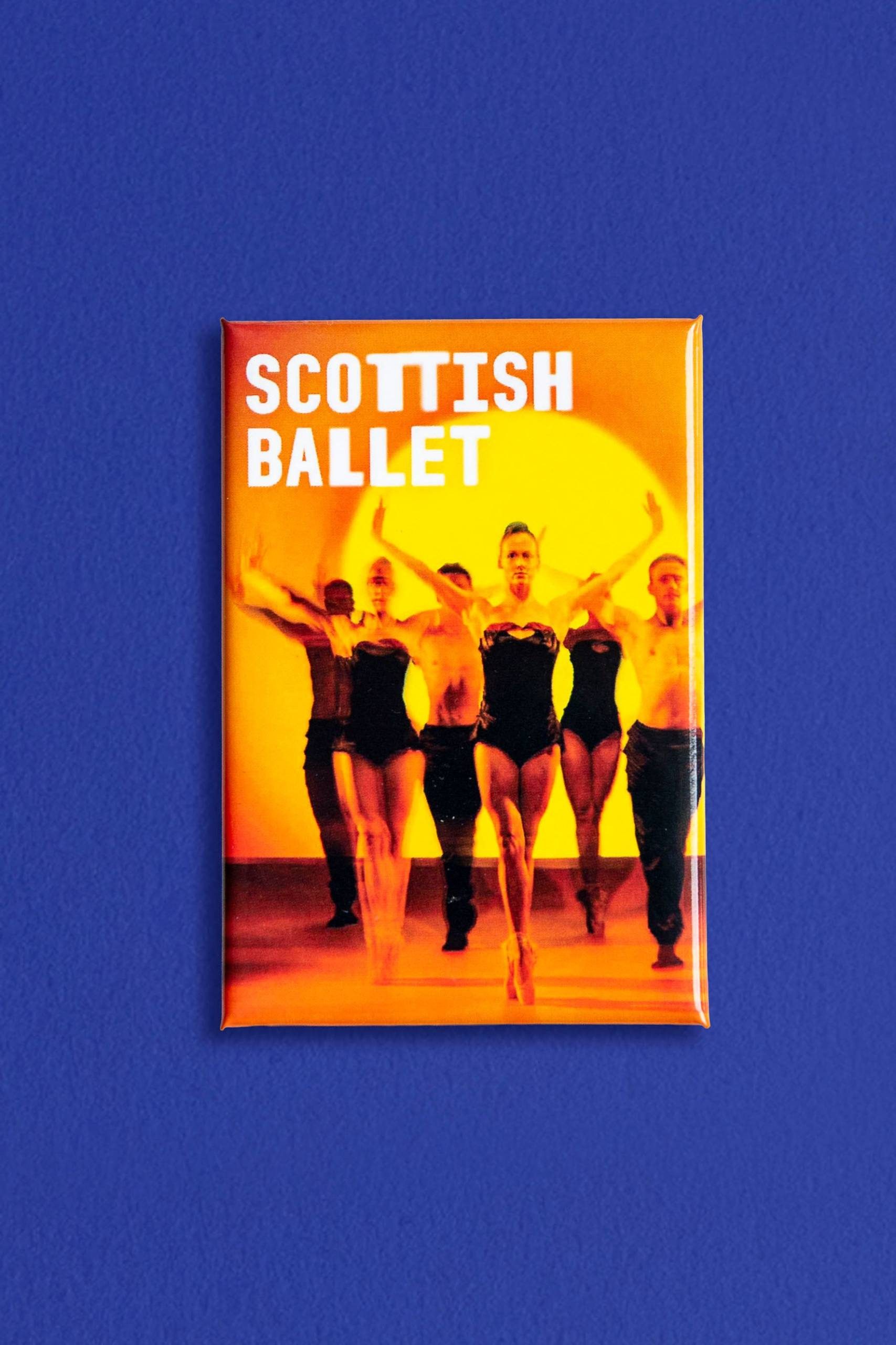A group of ballet dancers pose under orange lighting. The text SCOTTISH BALLET is displayed in bold white letters at the top. The image is set against a solid blue background.