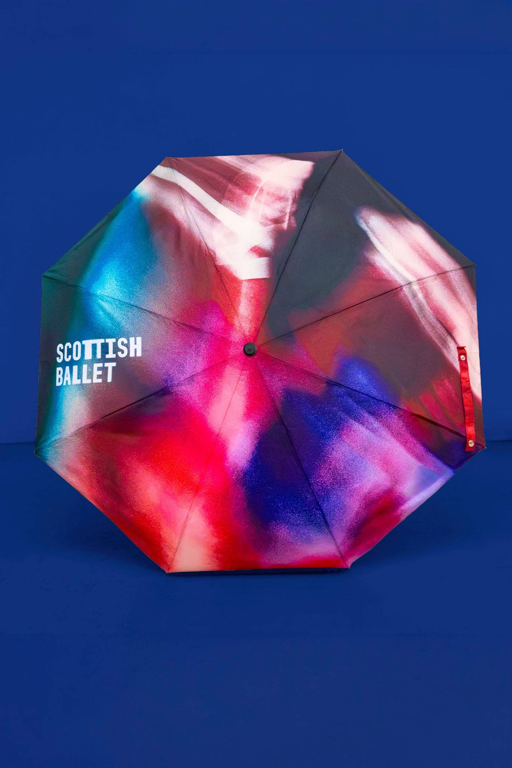 A colourful umbrella with abstract red, blue, and white patterns, featuring the text SCOTTISH BALLET on one panel, set against a solid blue background.