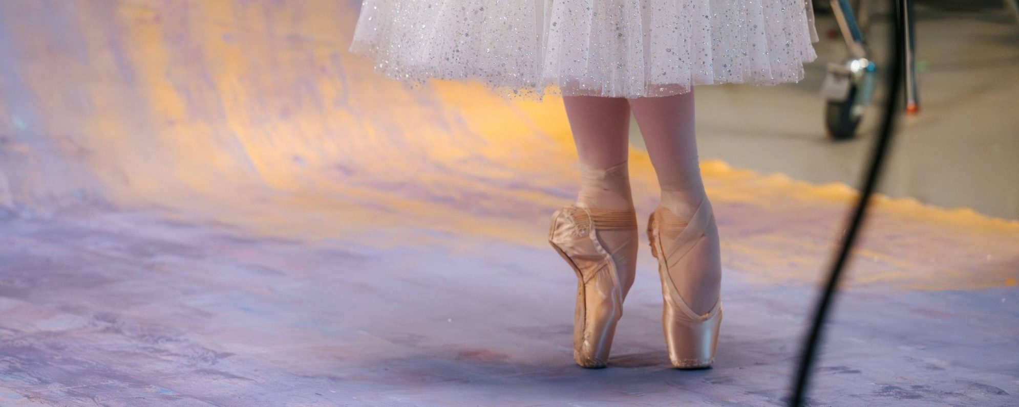 A ballerina stands en pointe on a purple and yellow painted floor, wearing a sparkly white tutu and ballet shoes. Only her lower legs and tutu are visible.