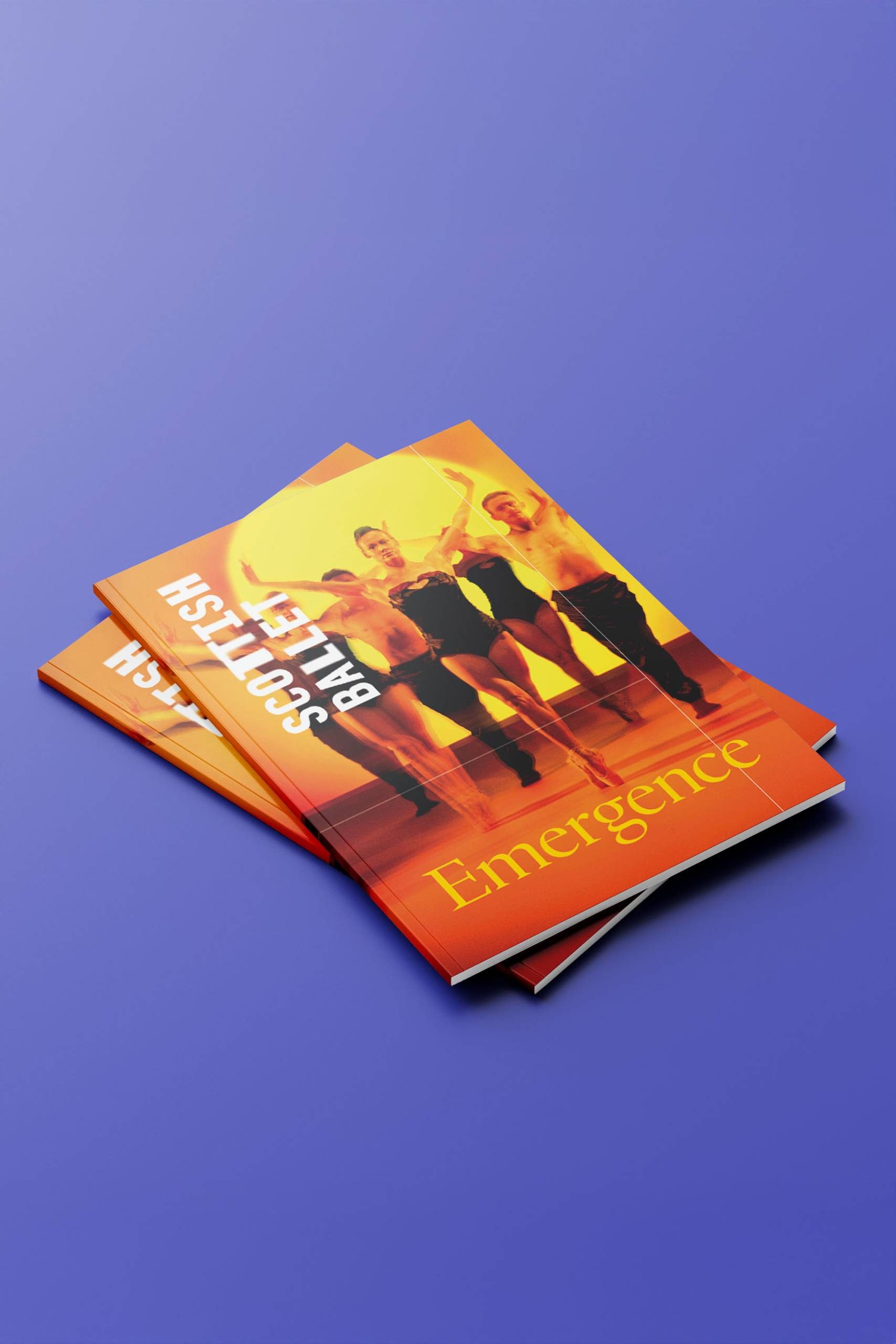 Two programmes on a purple surface show dancers on the cover under bright yellow-orange light. The text reads SCOTTISH BALLET at the top and Emergence at the bottom.