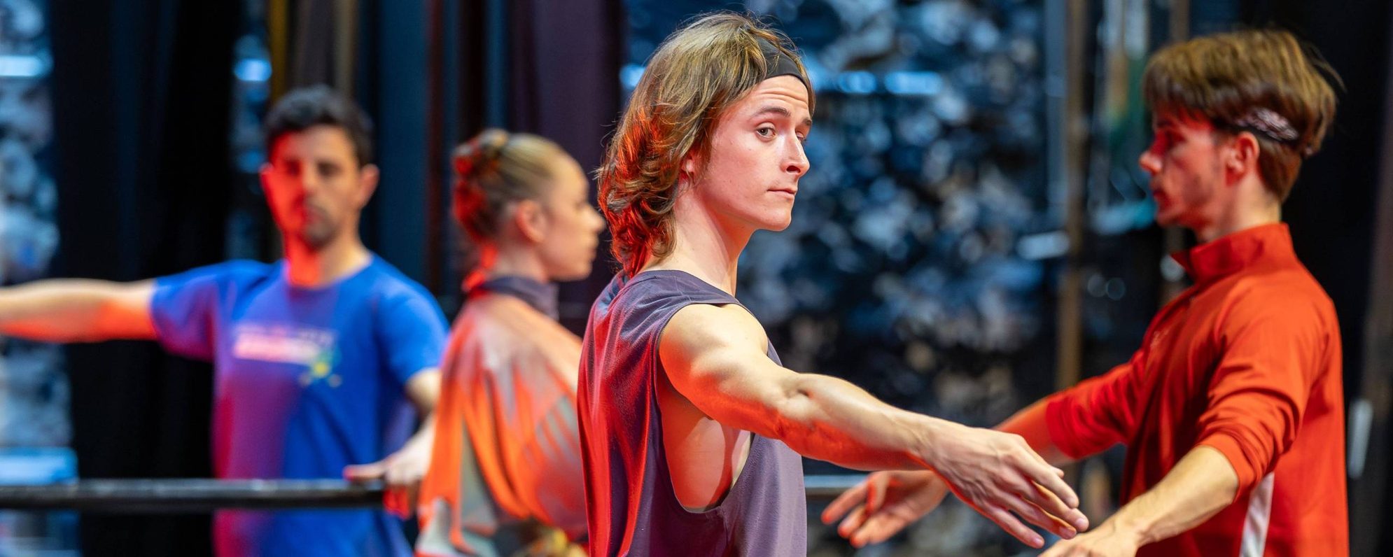 Four ballet dancers practise at a barre in a studio, focussing on their movements. The foreground dancer wears a sleeveless top and headband, extending their arms gracefully. The background shows blurred dancers and equipment.
